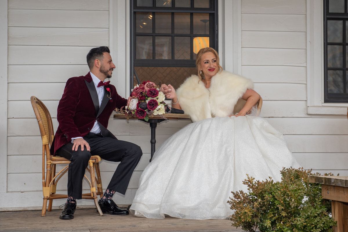 A bride in white and groom in velvet tuxedo share an intimate moment on a porch, hands touching over flowers.