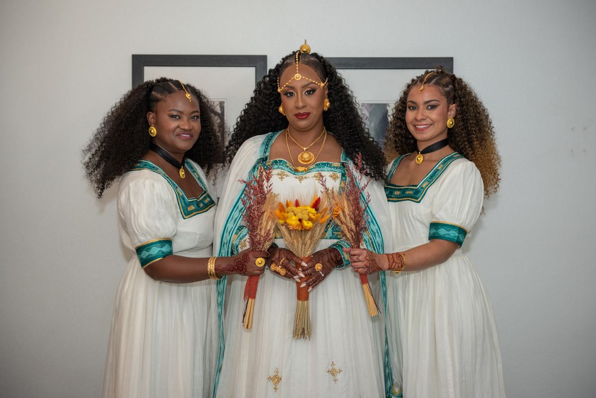 Three women in white and teal attire hold a dried floral arrangement, smiling against a neutral background with framed art.