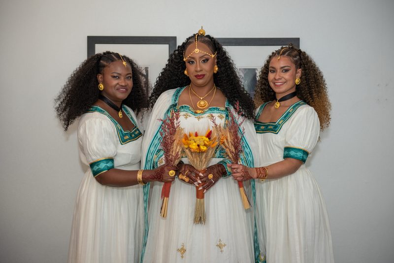 Three women in white and teal attire hold a dried floral arrangement, smiling against a neutral background with framed art.