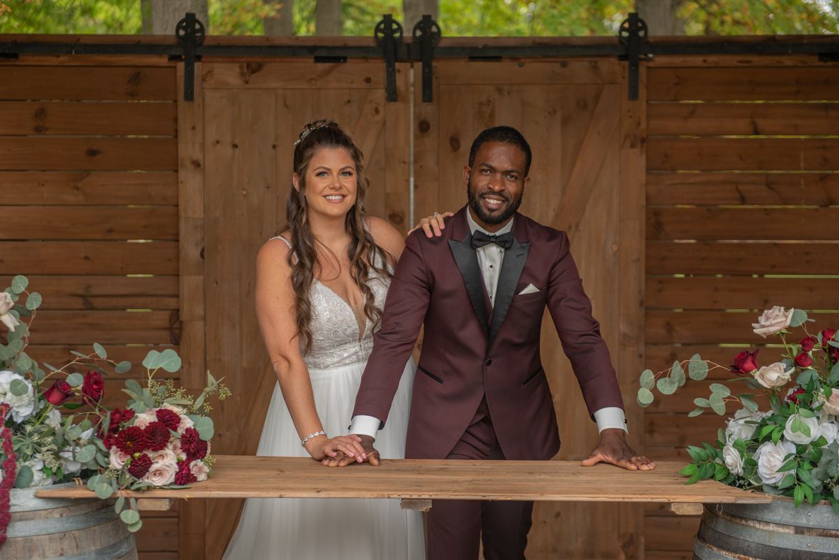 Alexandria and Derado stand side-by-side behind a rustic wooden table, smiling warmly at the camera.