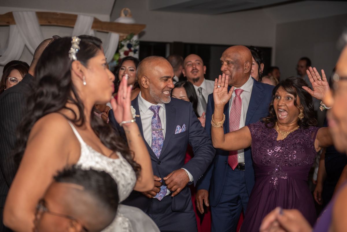 A bride in white and groom in navy suit laugh with guests at a reception surrounded by smiling family members and friends.