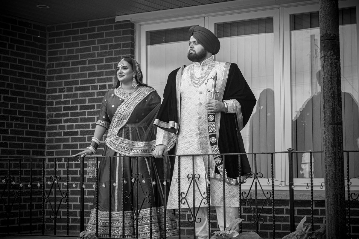 A bride and groom in traditional South Asian attire stand on a balcony, gazing outward with serene expressions.