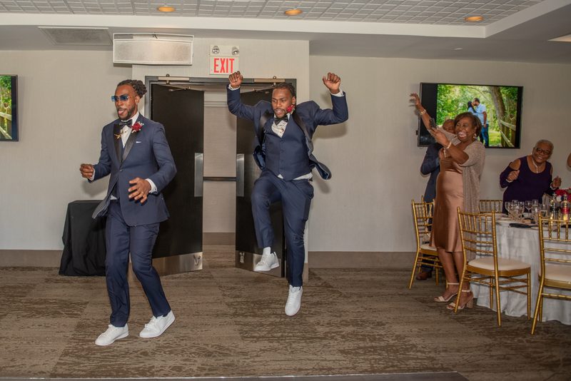Two men in navy suits dance energetically near an exit door as guests cheer, celebrating under bright lights.