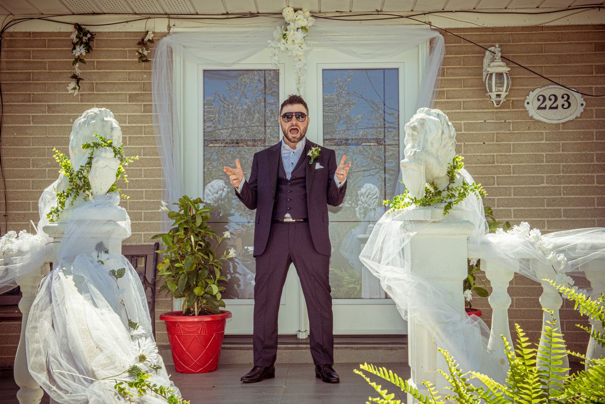 A man in a dark suit and sunglasses gestures dramatically on a porch flanked by veiled statues, with a festive brick house.