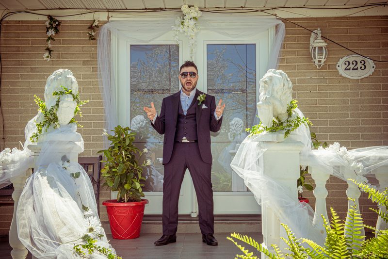 A man in a dark suit and sunglasses gestures dramatically on a porch flanked by veiled statues, with a festive brick house.