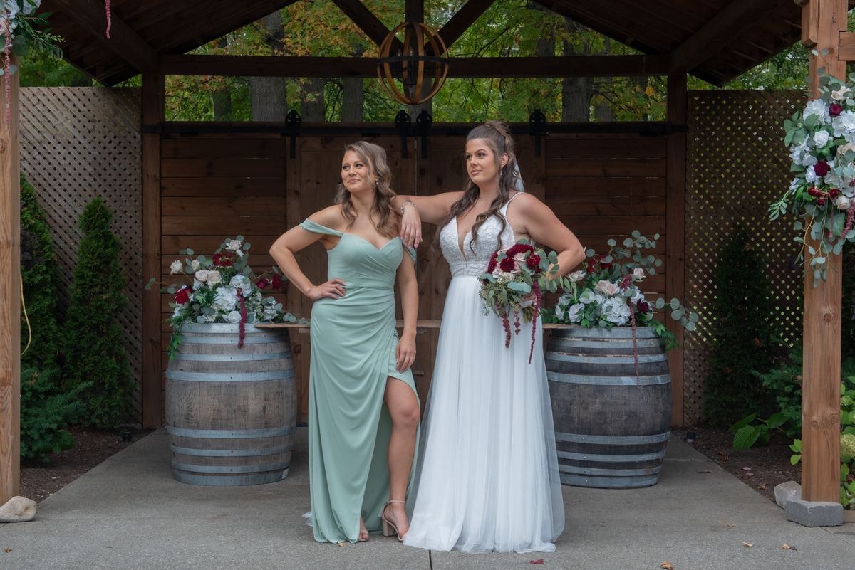Two women pose confidently under a wooden arbor adorned with floral arrangements on wine barrels, bathed in natural light.