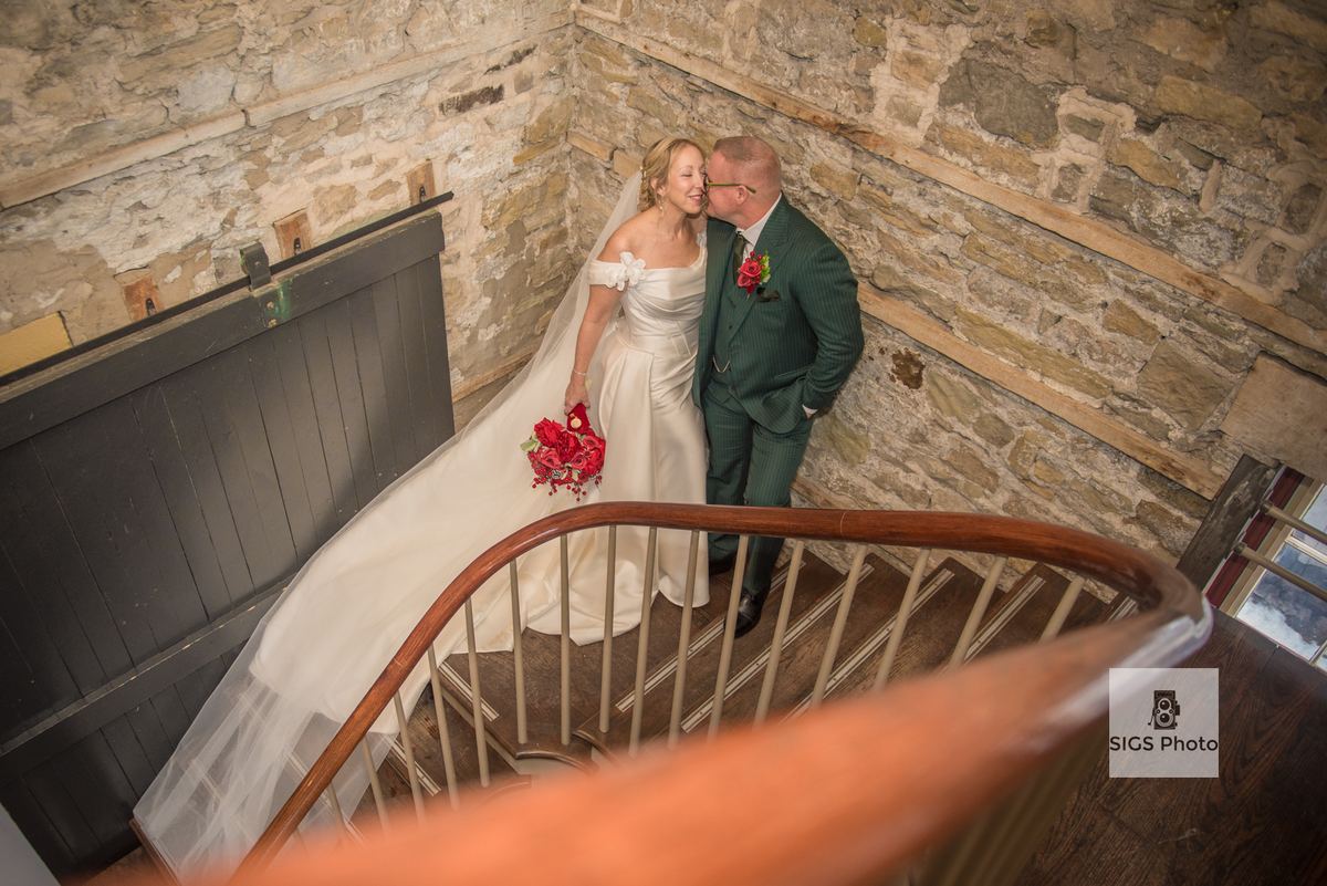 A bride and groom embrace on a wooden staircase inside a rustic stone building, natural light highlighting their joy.