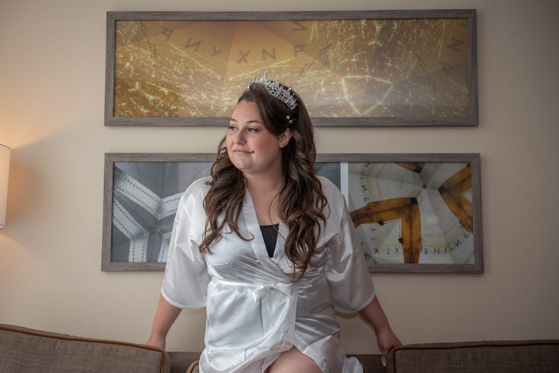 A bride in a white satin robe and tiara sits on a couch, gazing thoughtfully at an abstract geometric wall art.