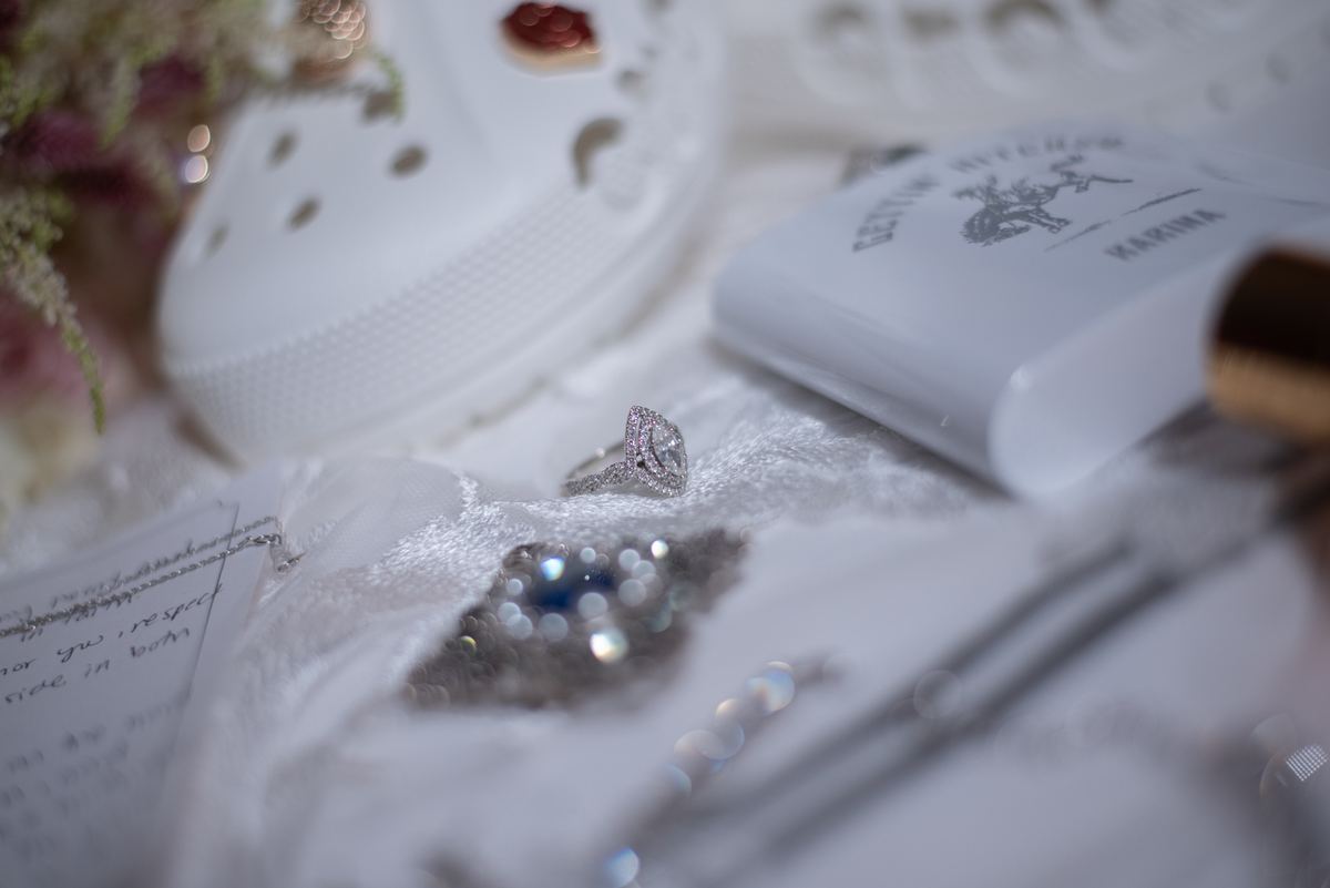 A sparkling engagement ring rests on white fabric with a note, box, and Crocs in soft lighting, evoking celebration.