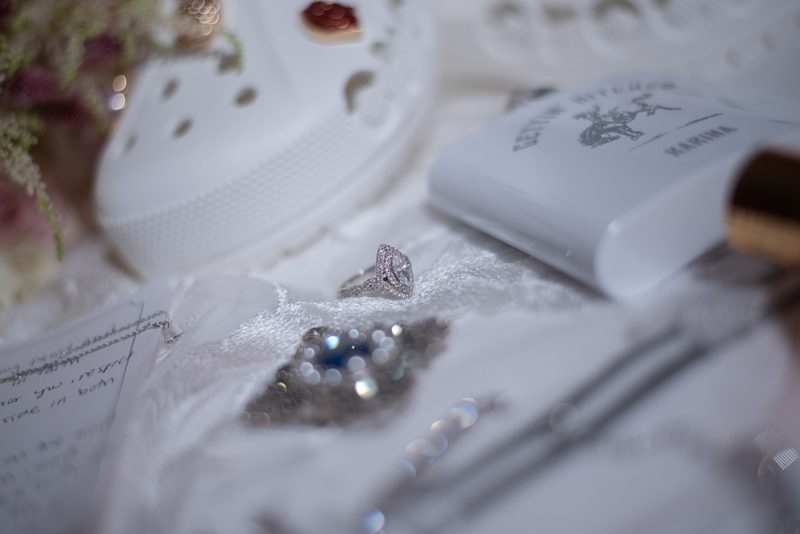 A sparkling engagement ring rests on white fabric with a note, box, and Crocs in soft lighting, evoking celebration.