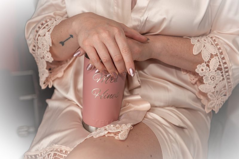 A bride in a soft pink satin robe with lace trim holds a personalized tumbler engraved with "Mrs. Hines".