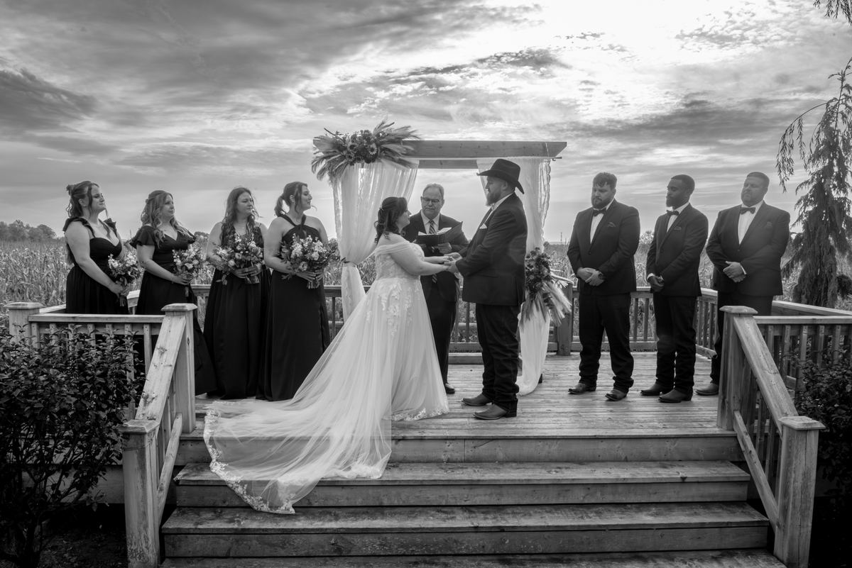 Bride and groom exchange vows under a floral arch on a wooden deck, surrounded by wedding party and officiant.