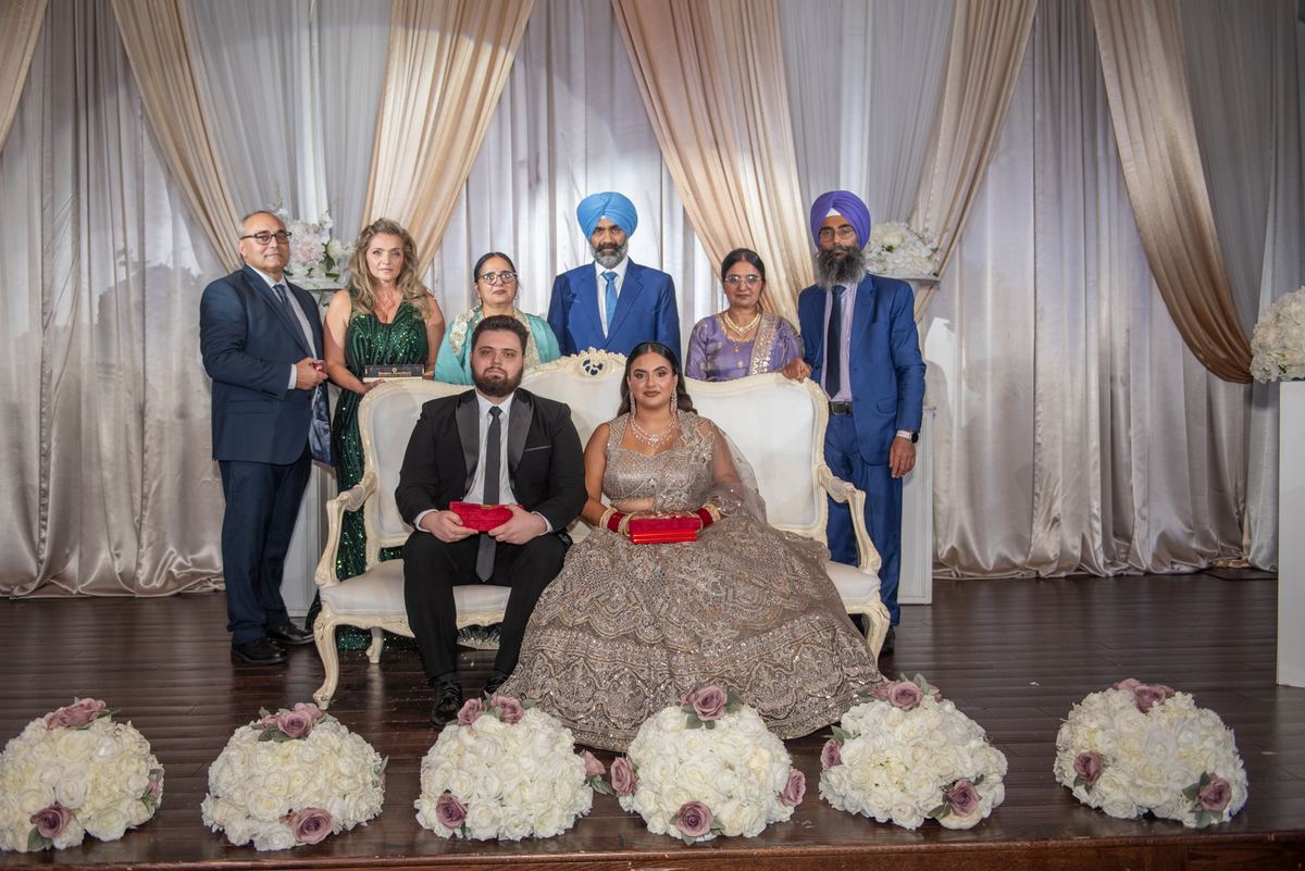 A bride and groom in formal attire sit with a group of formally dressed guests behind them, illuminated by stage lights.