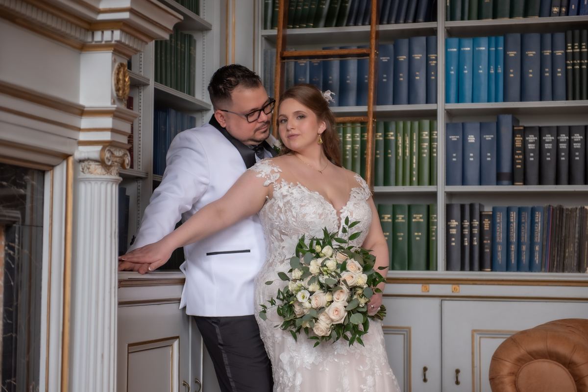 A bride and groom stand in an elegant library, the groom leaning close as she holds a bouquet, surrounded by soft lighting.