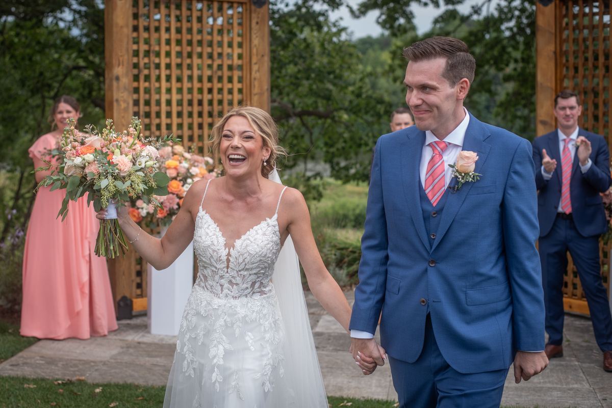 A laughing bride walks down an outdoor aisle with her groom, surrounded by guests and wooden lattice.