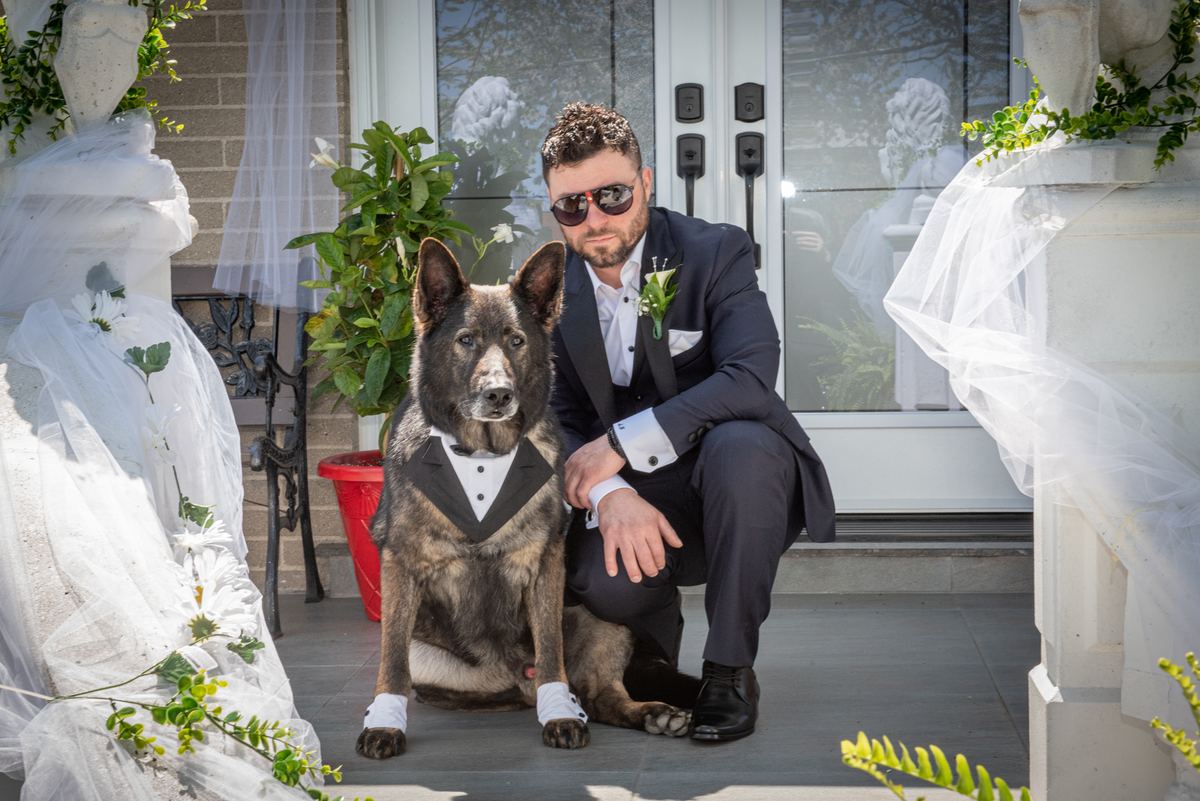 A groom in tuxedo and sunglasses poses with his miniature dog dressed as a tuxedoed groom on a decorated porch.