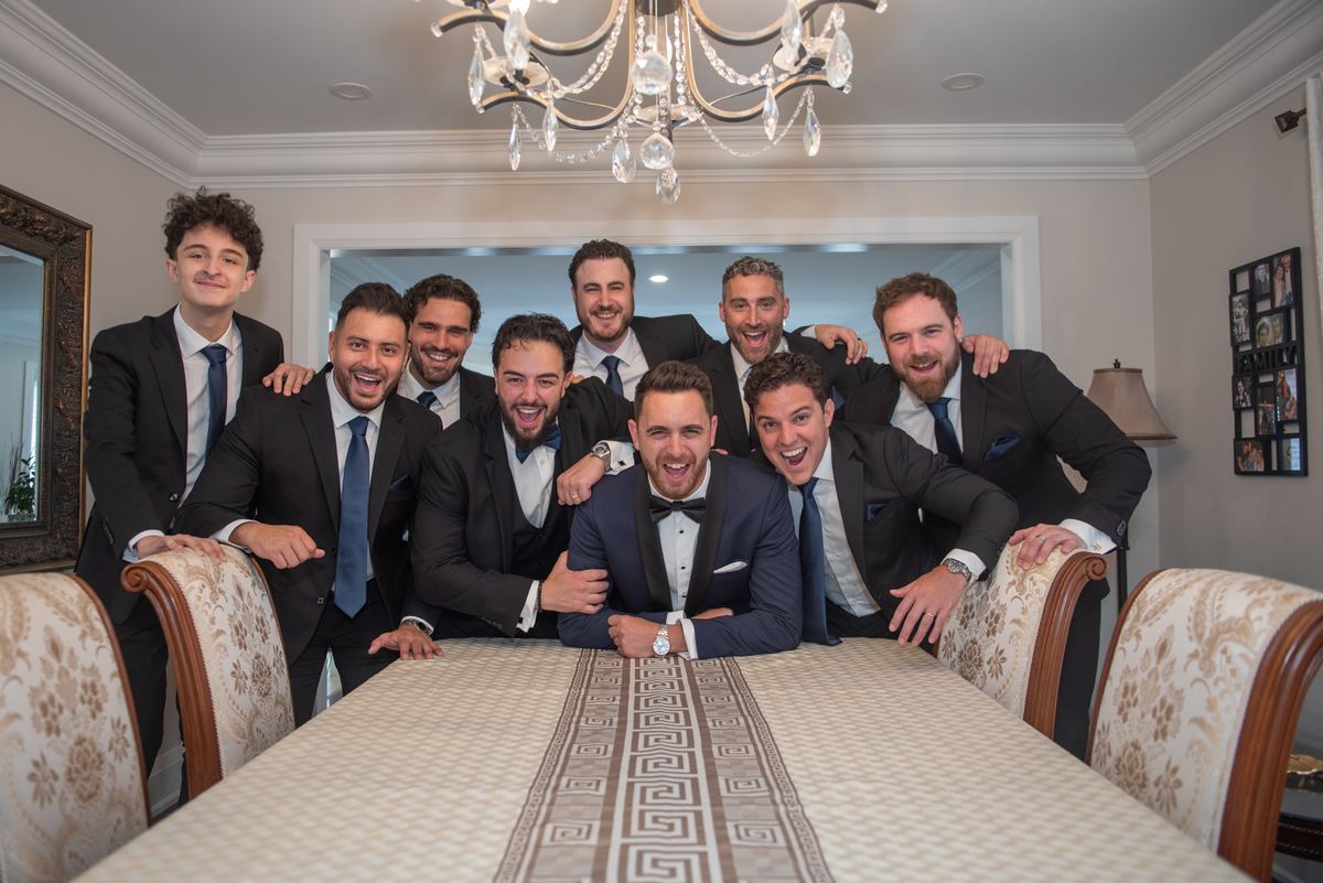 Groom and his smiling groomsmen pose in a formal dining room around a decorated table, illuminated by an ornate chandelier.