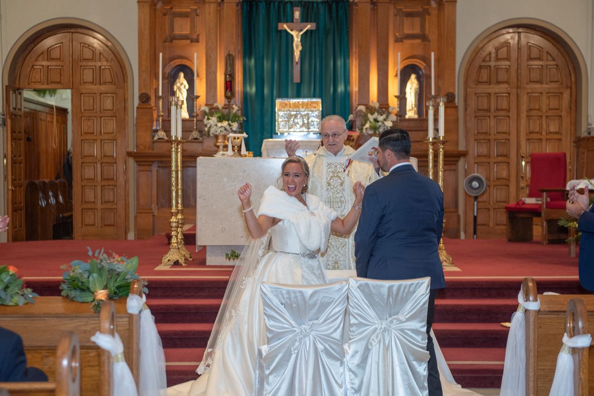 A bride in white reacts emotionally beside her groom during a church ceremony with a priest and floral decorations.