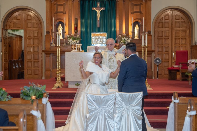 A bride in white reacts emotionally beside her groom during a church ceremony with a priest and floral decorations.