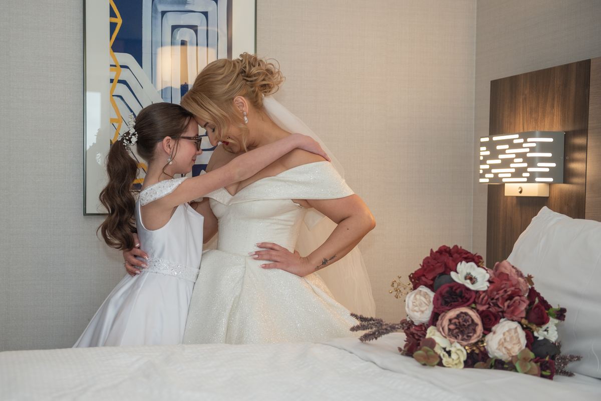 Bride in off-shoulder gown embraces young girl in white dress, their foreheads touching near wall sconce.