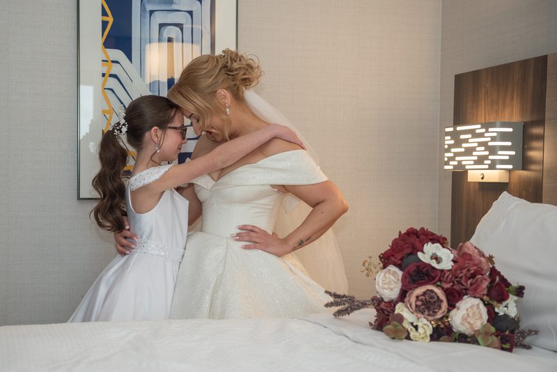 Bride in off-shoulder gown embraces young girl in white dress, their foreheads touching near wall sconce.