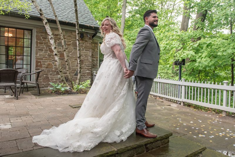 A bride and groom stand on a stone patio, the bride laughing in her lace gown as confetti dots the ground.