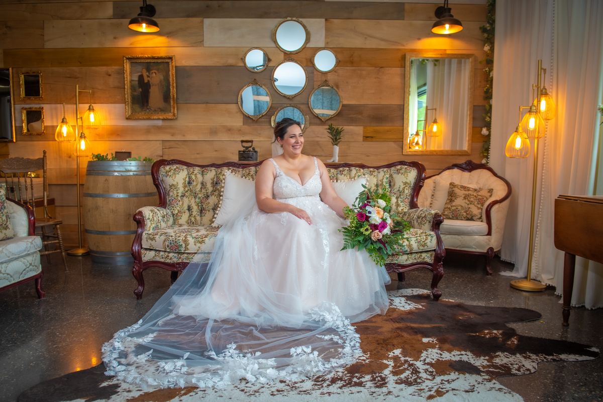 A smiling bride in a white wedding gown sits on a floral sofa in a rustic room with wooden walls and golden lighting.
