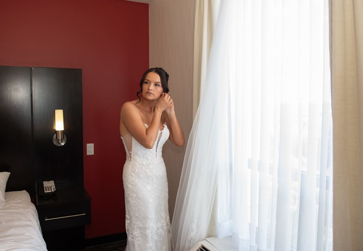 A bride in a white lace gown adjusts her veil near a window, bathed in soft daylight and the glow of a wall sconce.