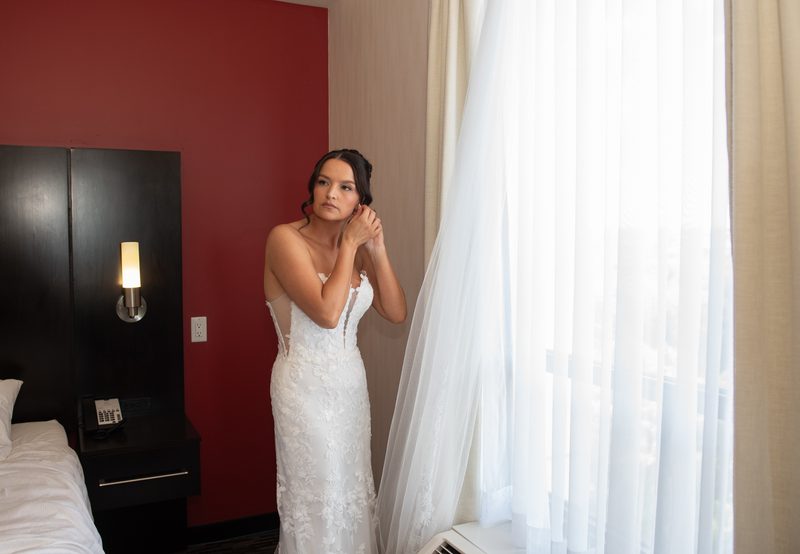 A bride in a white lace gown adjusts her veil near a window, bathed in soft daylight and the glow of a wall sconce.