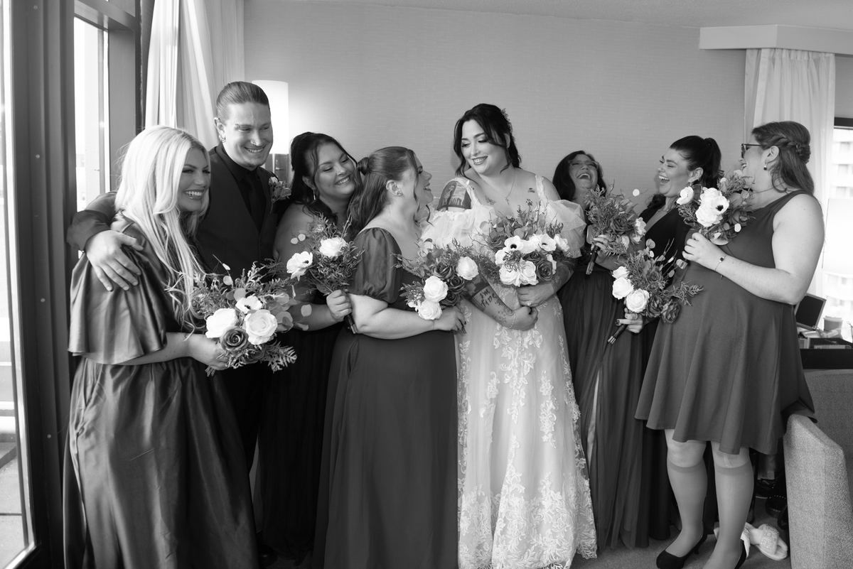 A bride and groom laugh with bridesmaids near a window, holding bouquets in a warmly lit room, emphasizing joy.