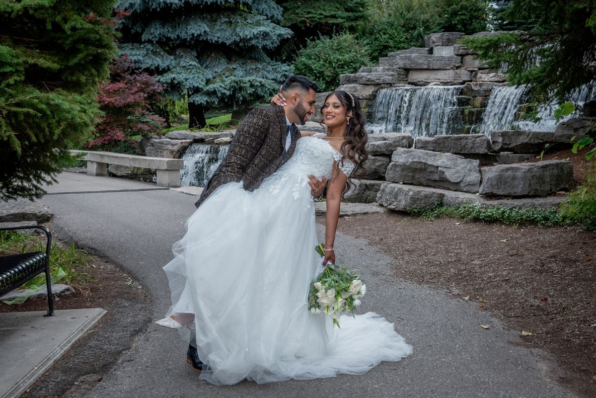 A bride in white and groom by a stone waterfall in a garden, sharing an intimate moment under diffused sunlight.