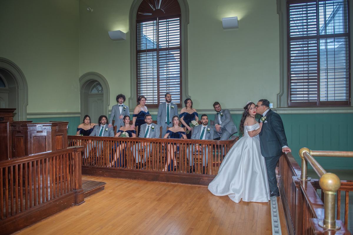 Bride and groom share an intimate moment by a railing in a grand, wood-paneled room with their wedding party behind them.