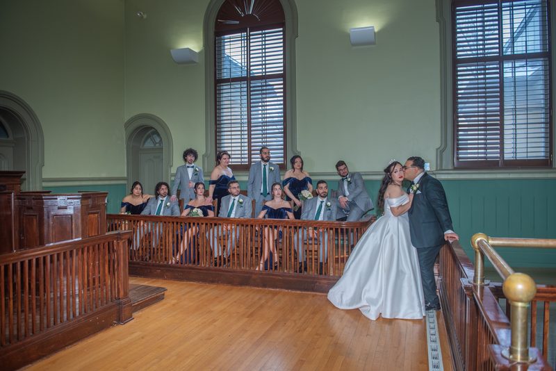 Bride and groom share an intimate moment by a railing in a grand, wood-paneled room with their wedding party behind them.