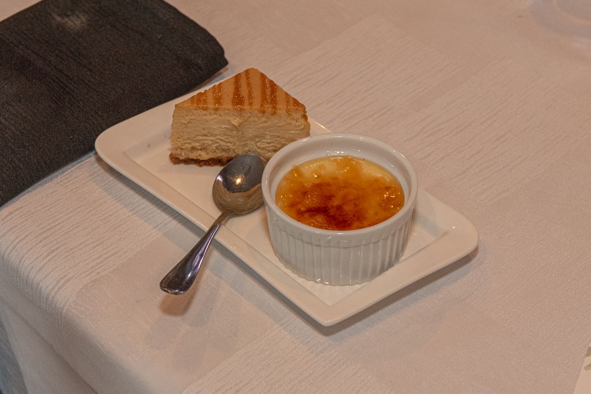 A white plate holds a slice of grilled cheesecake and crème brûlée with a spoon nearby, under soft lighting.