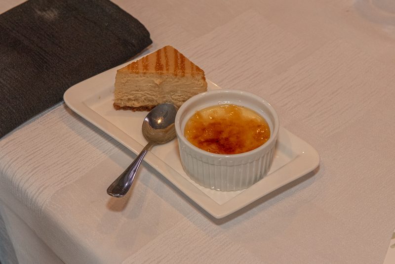 A white plate holds a slice of grilled cheesecake and crème brûlée with a spoon nearby, under soft lighting.