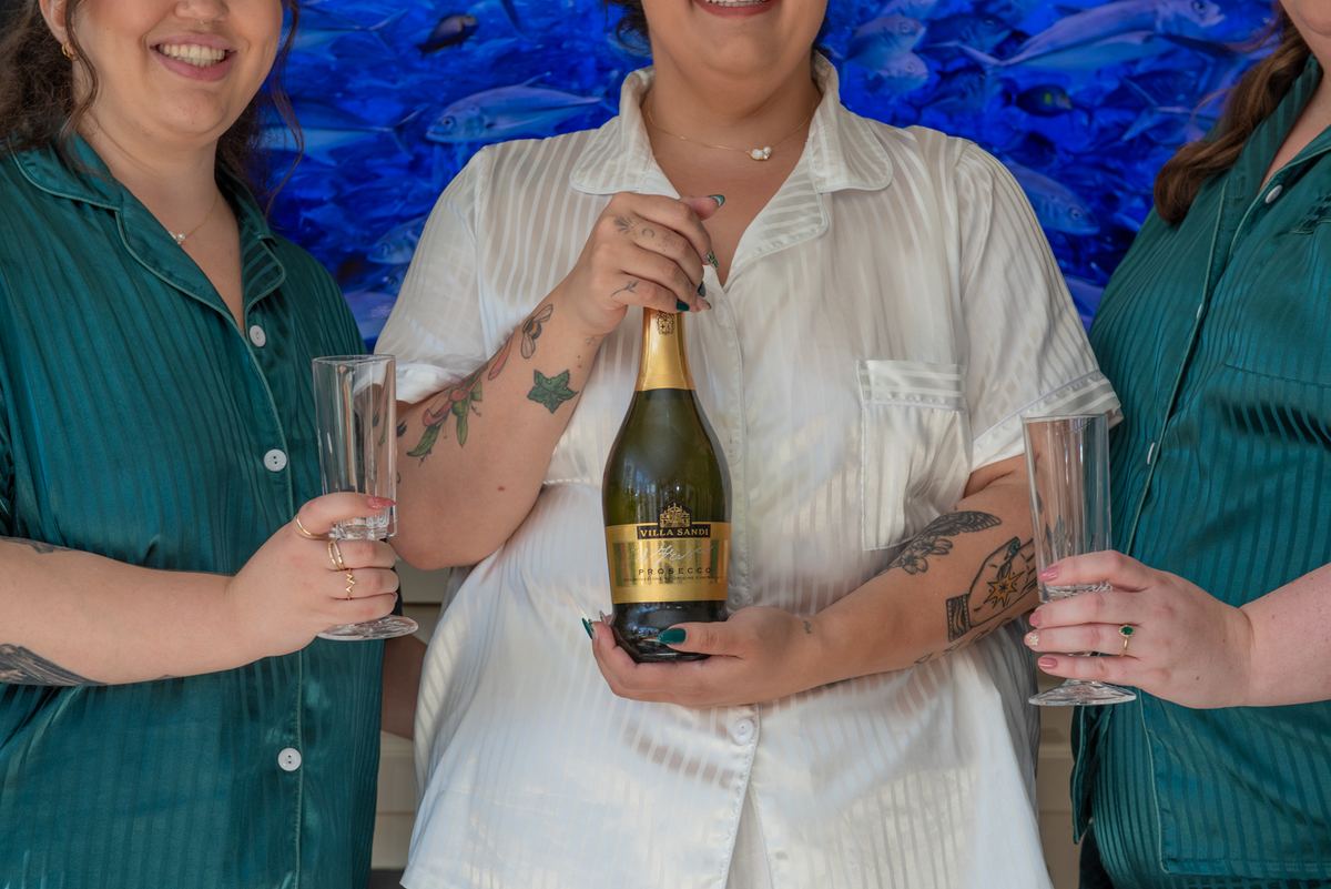 Three women smiling around a bottle of Prosecco, one holding the bottle against a blue fish-patterned backdrop.