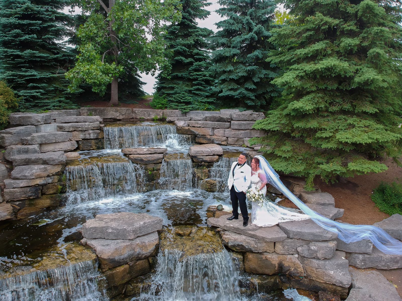 A newlywed couple stands beside a waterfall with lush evergreen trees in the background, the bride's veil flowing behind her.