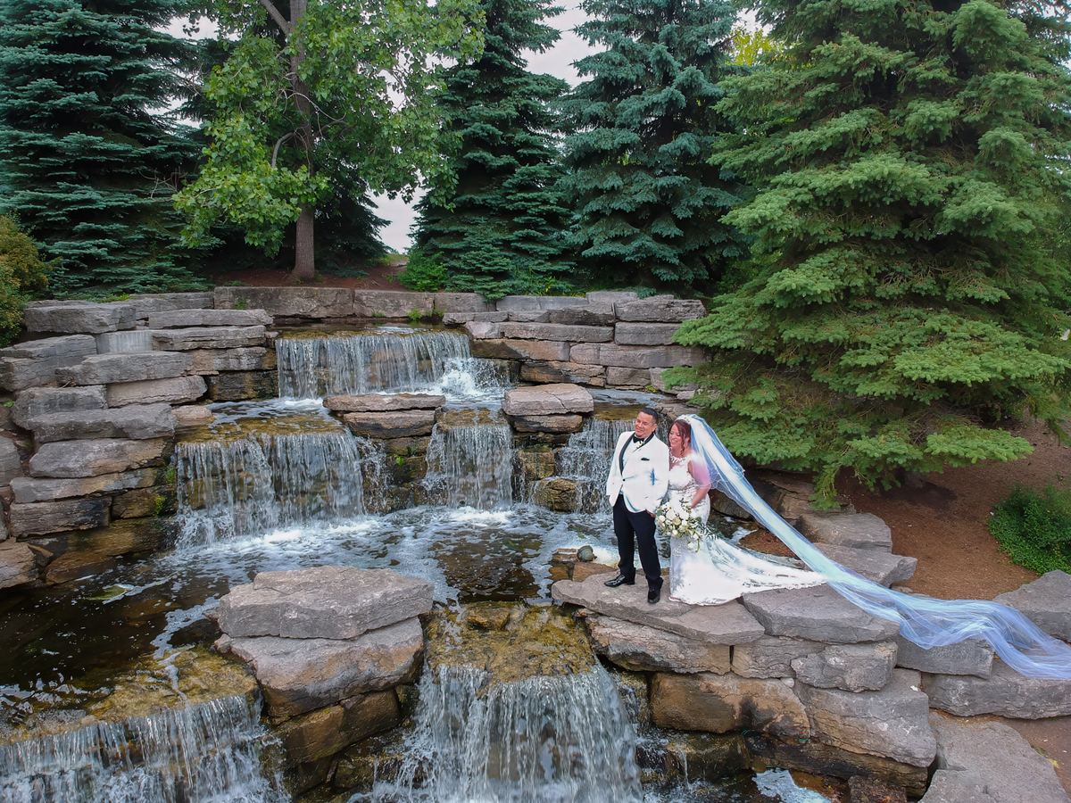 A newlywed couple stands beside a waterfall with lush evergreen trees in the background, the bride's veil flowing behind her.