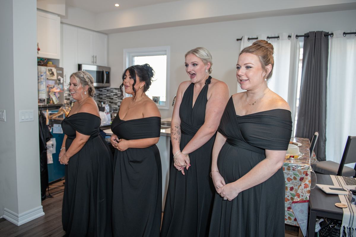 Four women in black gowns stand in a well-lit room, reacting with surprise and delight to an unseen event.