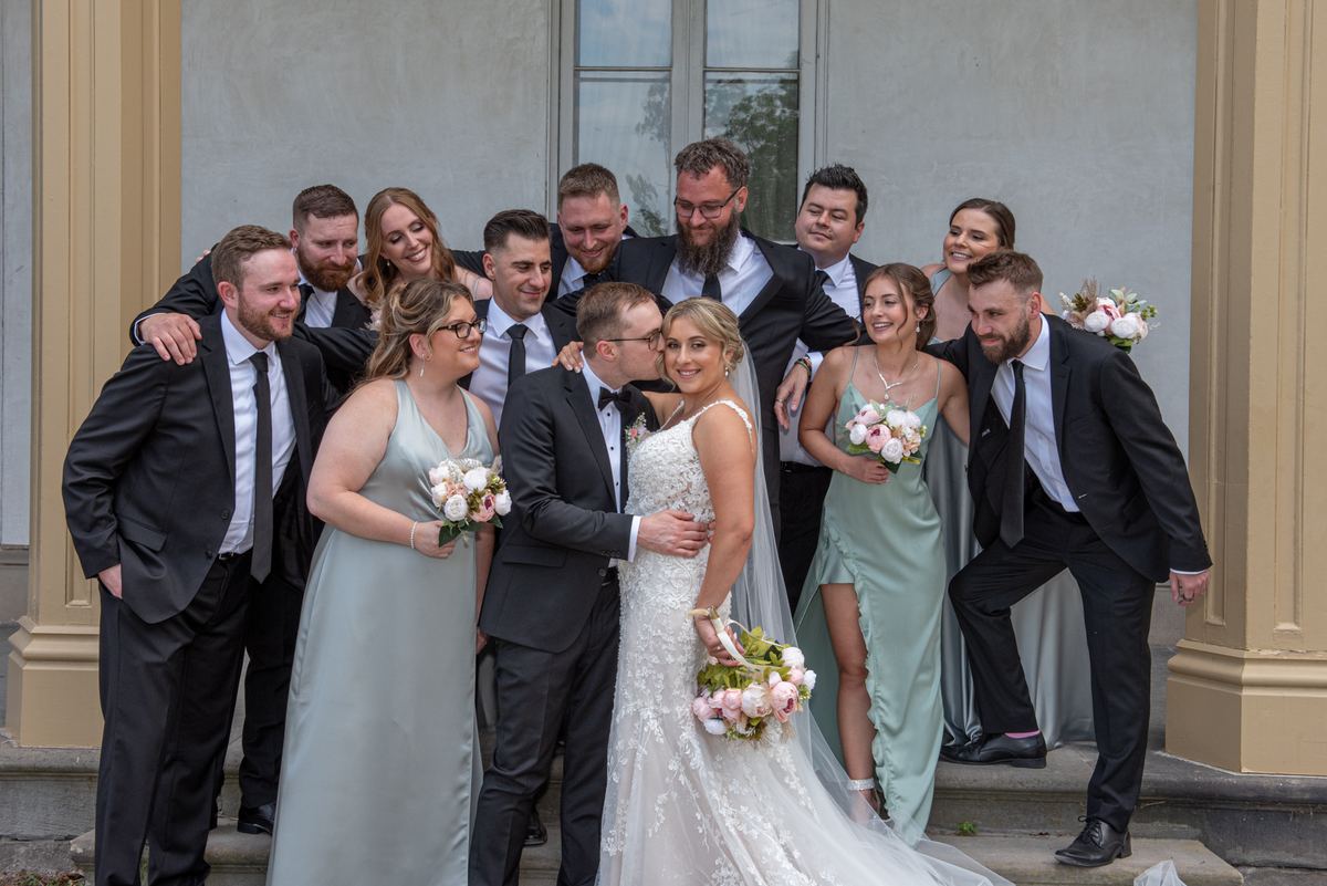 A bride and groom embrace on steps with their smiling wedding party in the background, illuminated by natural light.