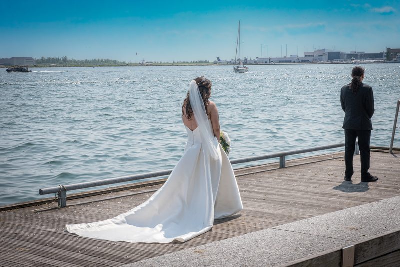 A bride in a white gown and veil stands on a wooden pier, gazing at the water with her groom nearby.