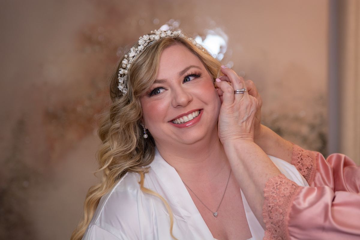 A bride smiles as a family member adjusts her hair beside her during wedding preparations, softly lit with warm light.