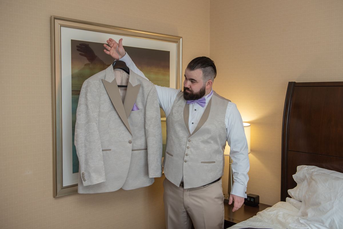 A groom in a light-colored tuxedo with a purple bow tie holds up his jacket near an unmade bed and framed artwork.