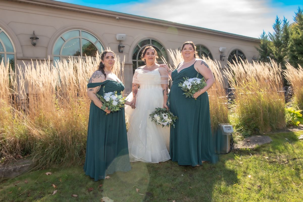 Three women, including a bride in a white gown, stand beside tall grasses holding bouquets of flowers and greenery.