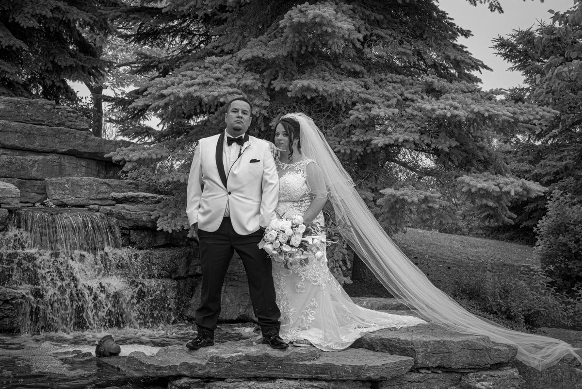 A bride in a lace gown and veil with a bouquet stands beside a stone waterfall with her groom in a tuxedo.