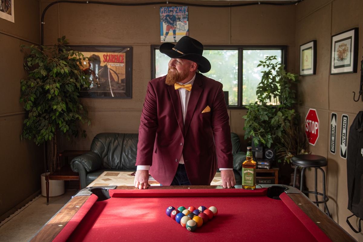 A man in a maroon suit and yellow bow tie stands by a pool table, gazing upward as light streams through the window.