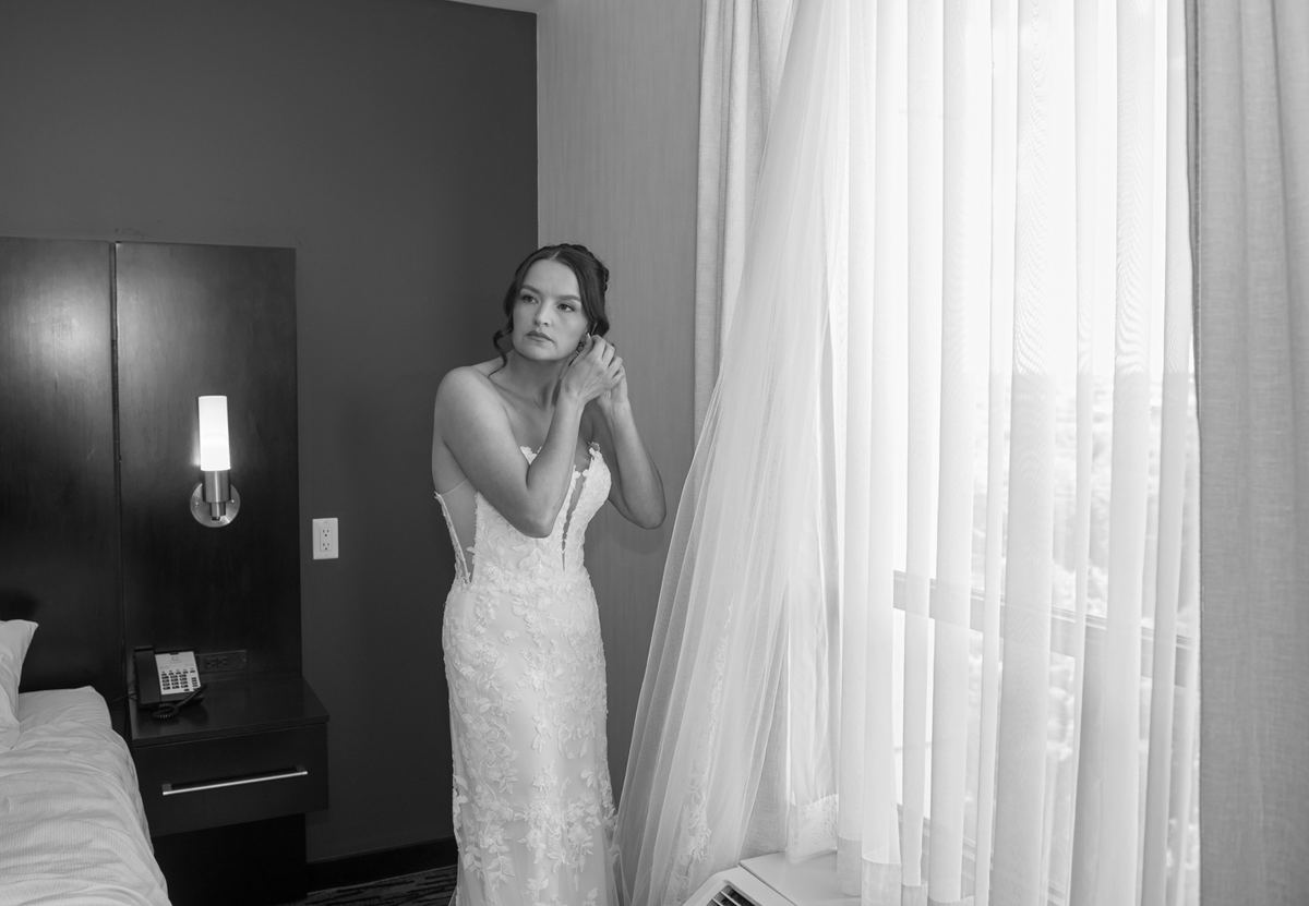 A bride in a wedding gown stands by a window, adjusting her veil thoughtfully as light highlights her face and dress.