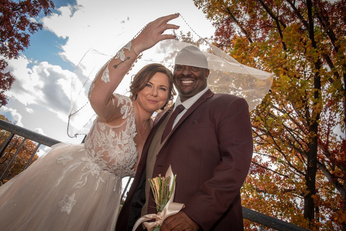 A bride in a lace gown and groom in a burgundy suit pose under autumn leaves with the bride lifting her veil.