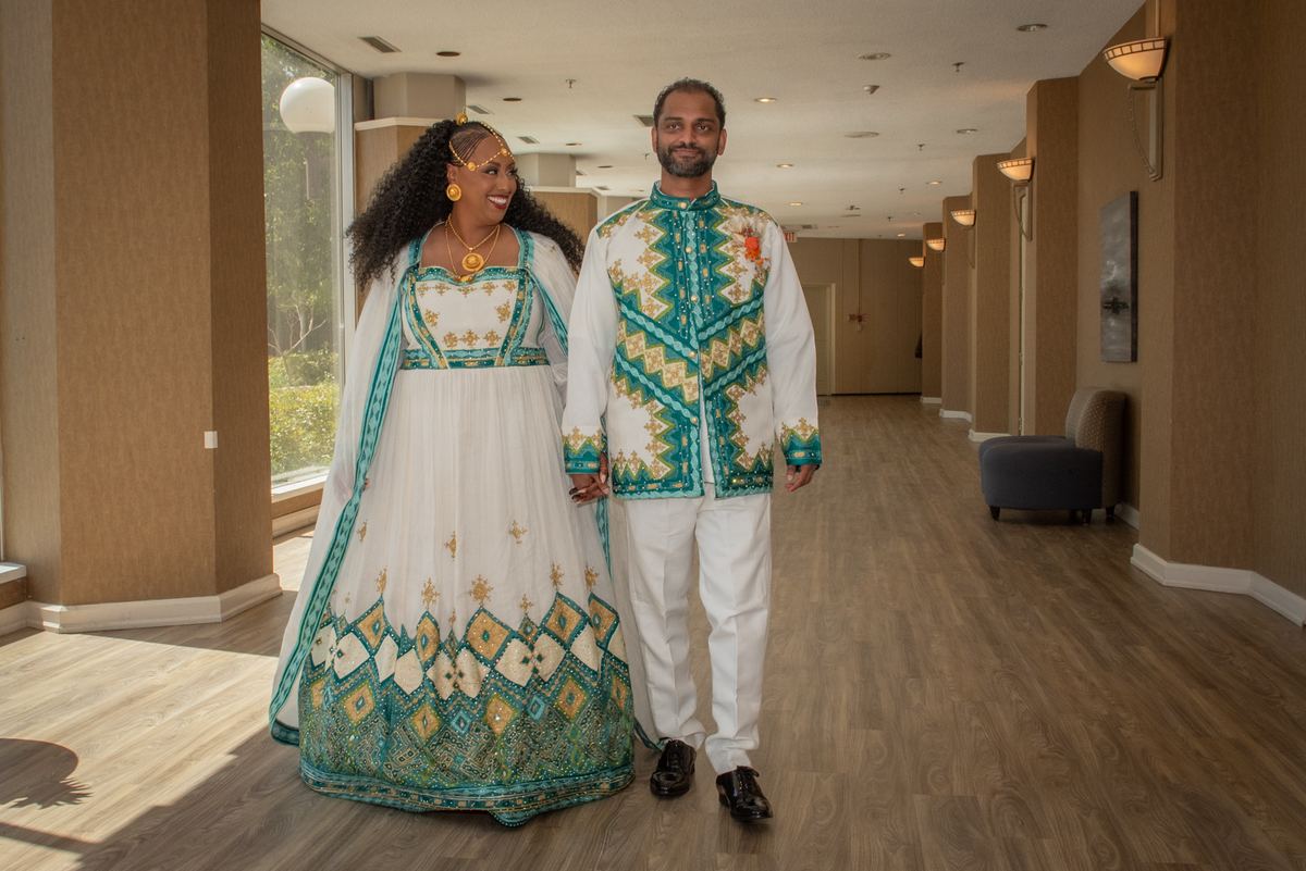 A smiling couple in teal and gold traditional attire walks down a lit hotel corridor, illuminated by natural light.