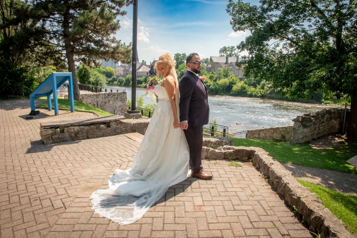 A smiling bride and groom stand hand-in-hand on a stone riverside walkway, framed by lush trees and a cityscape.